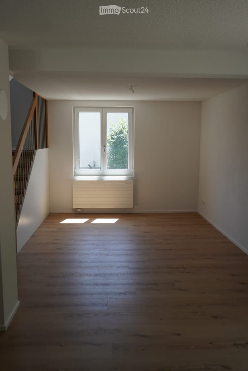 Empty room with wooden flooring, white walls, a window with white frame, and a radiator below it. The room is well-lit with natural light coming through the window.