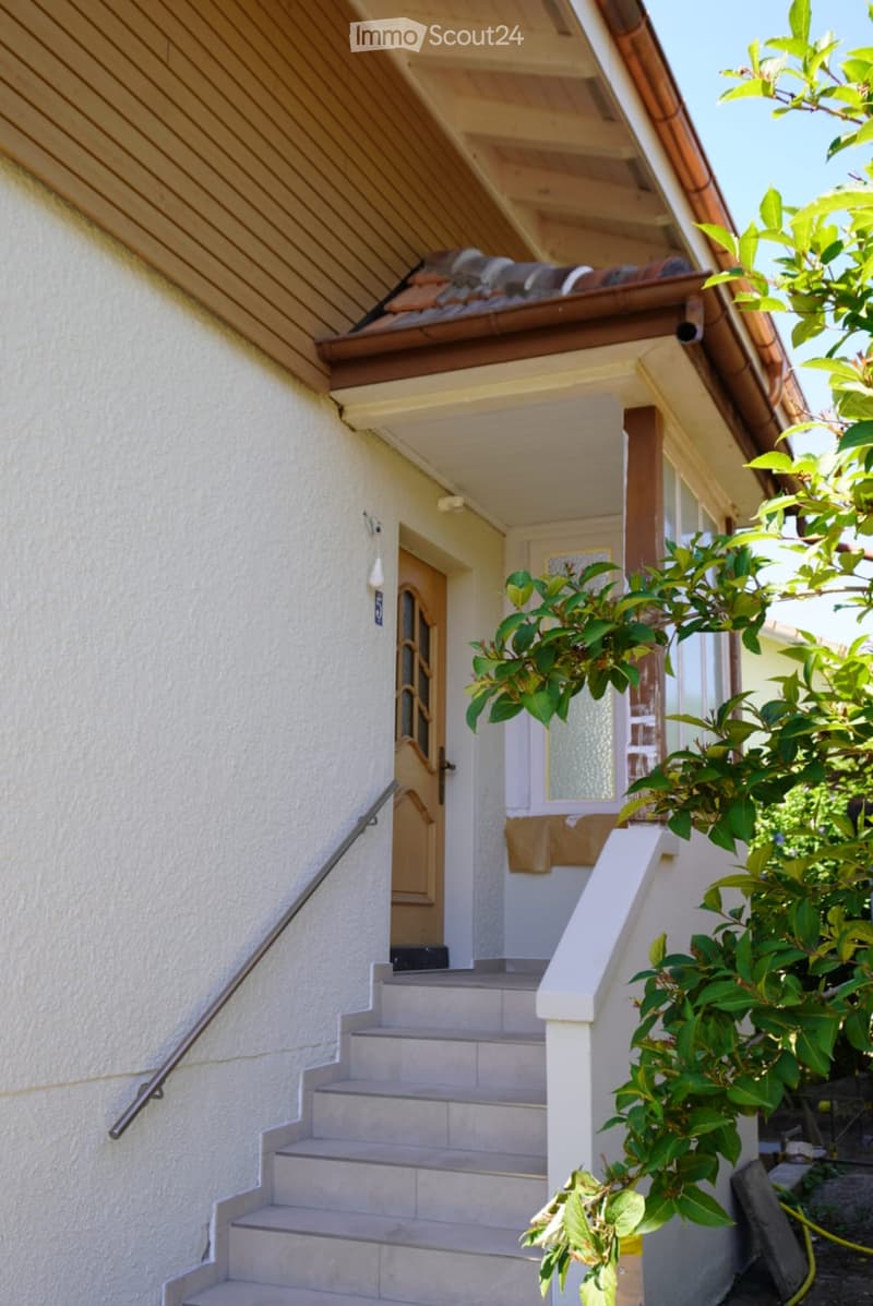 Exterior view of a house with a raised ground floor, featuring a staircase leading to the entrance, a covered porch area, and greenery on the right side.