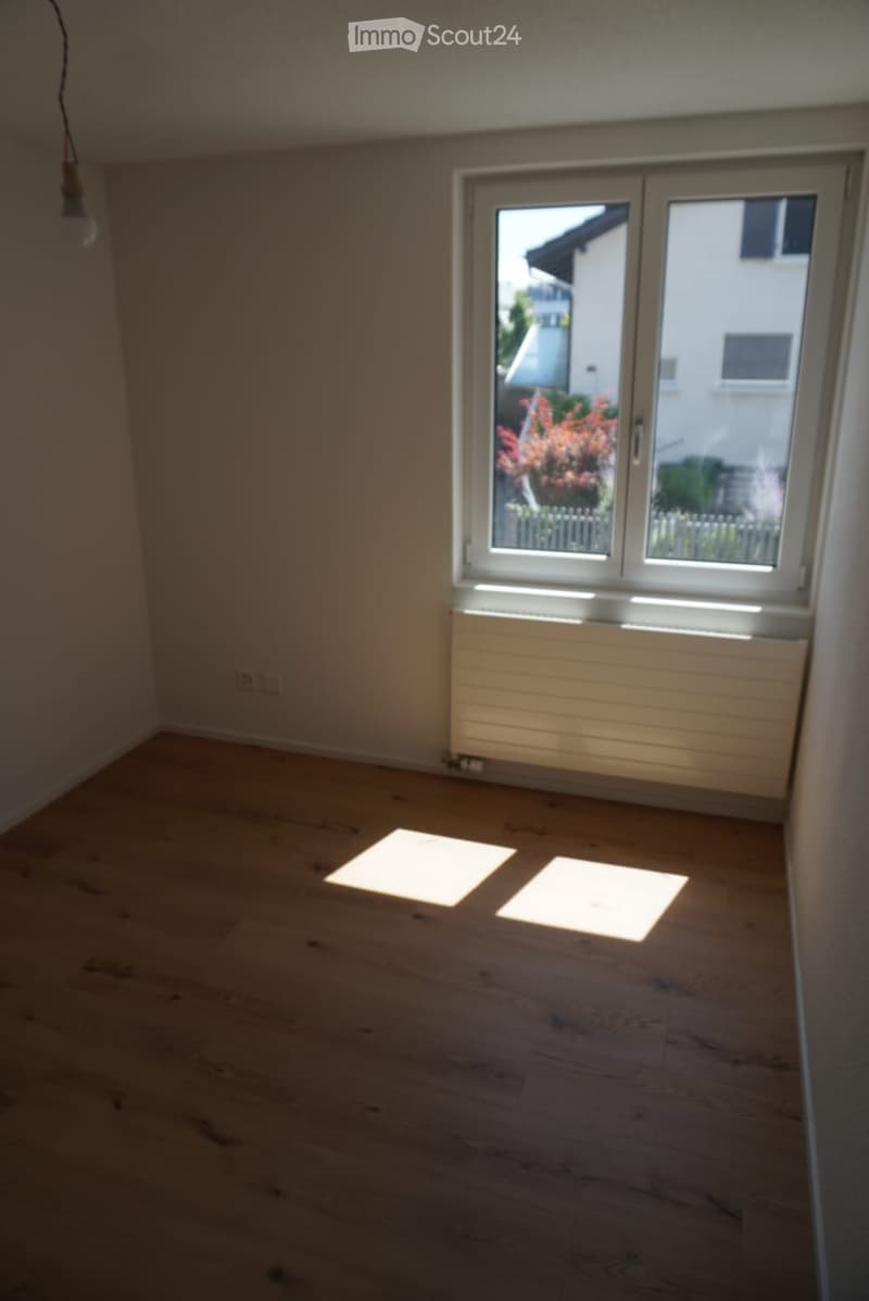 Empty room with wooden flooring, white walls, and a large window with a view of a garden and neighboring building.