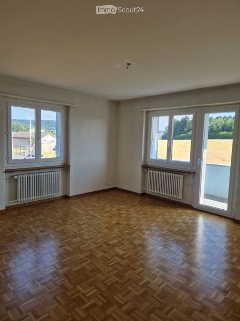 Empty room with wooden parquet flooring, white walls, large windows, radiators, and natural light.