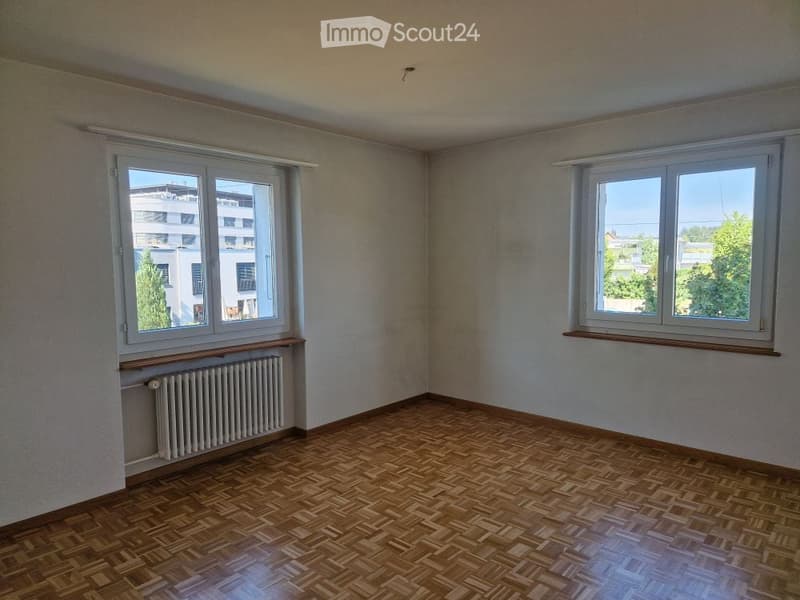 Empty room with wooden parquet flooring, light-colored walls, and two windows with white frames. Radiator visible under one window.