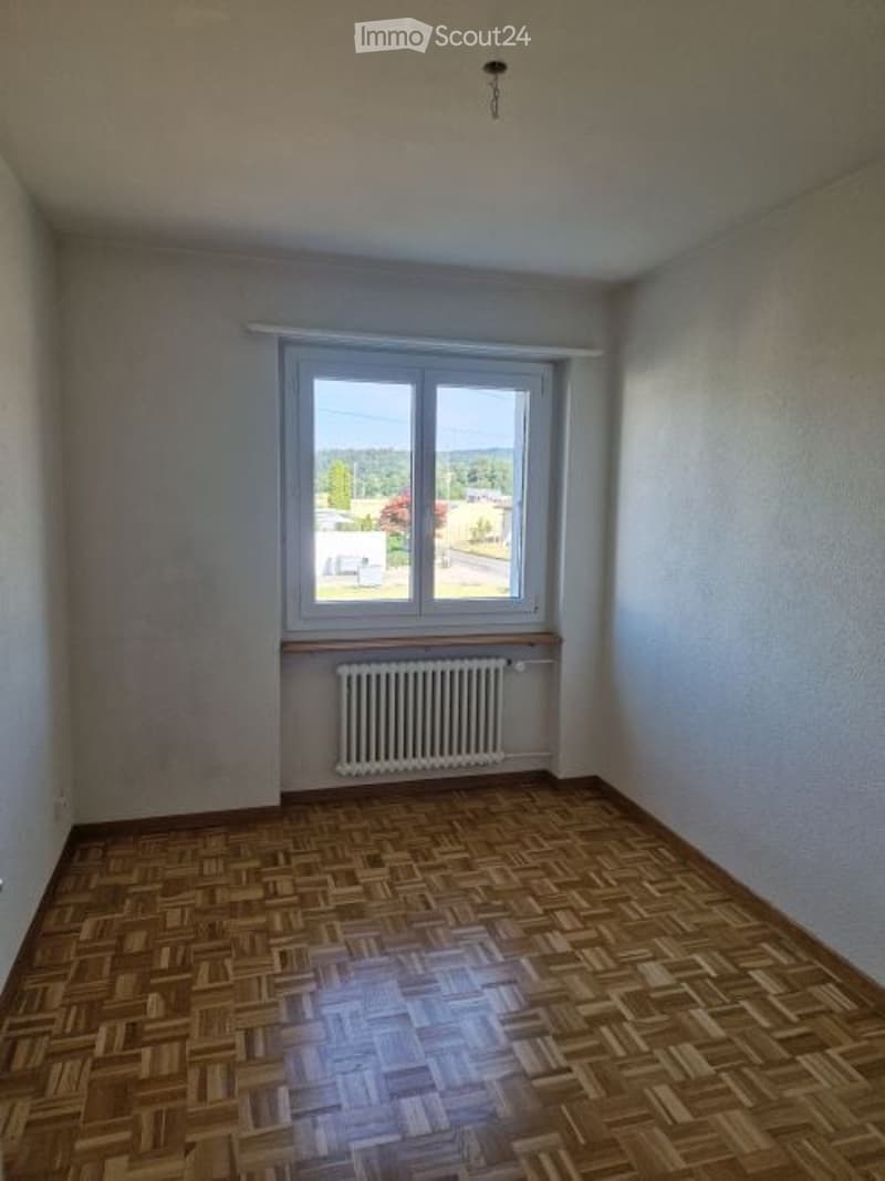 Empty room with wooden parquet flooring, white walls, a single window with white frame, and a radiator below the window.