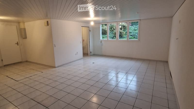 Empty room with tiled floor, white walls, and large windows showing greenery outside. No furniture or equipment present.