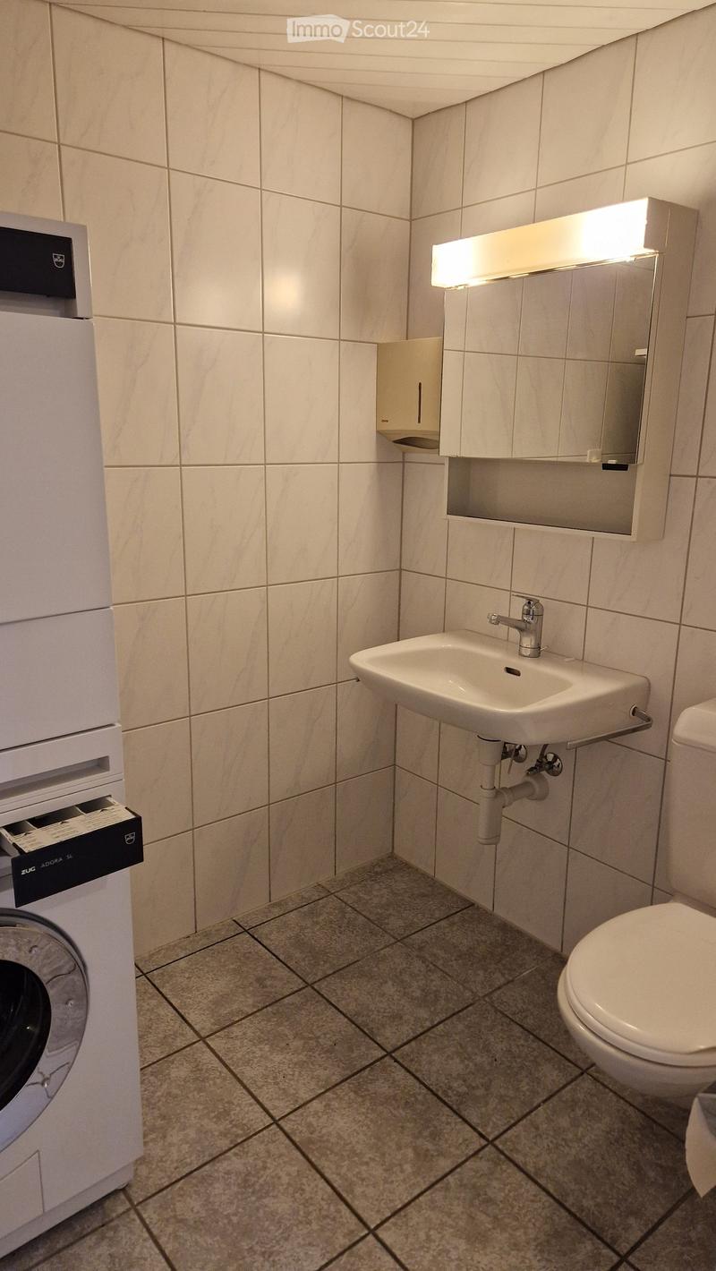 Modern bathroom with white tiled walls, a sink with a chrome faucet, a toilet, a mirror with built-in lighting, and a washing machine.