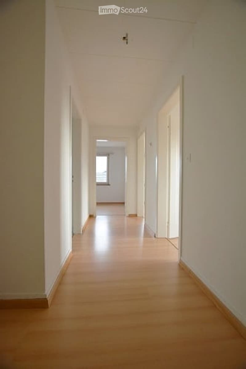 Empty hallway with light wooden flooring and white walls, leading to a room with a window at the end.