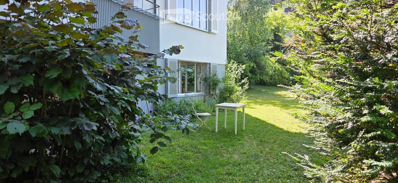A well-maintained garden with a small white table and a folding chair, surrounded by lush greenery and a white building with large windows in the background.
