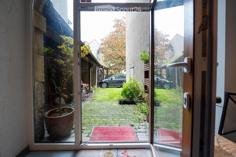 View from inside a house through an open door to a small, paved garden area with potted plants, a red doormat, and a grassy lawn. Two parked cars are visible in the background.