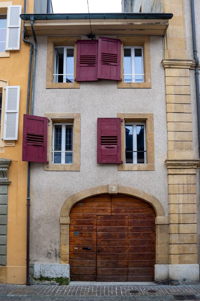 Facade of a two-story building with a wooden garage door, multiple windows with red shutters, and a textured beige wall.