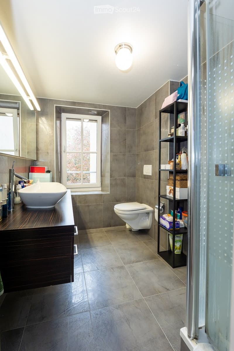 Modern bathroom with grey tiles, a white sink, a toilet, a shelf with various items, and a large window providing natural light.