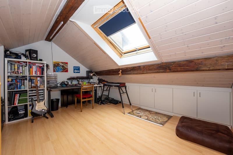 Bright attic room with wooden floors, slanted wooden ceiling, skylight window, desk with chair, bookshelf, guitar, cabinet, and a small keyboard setup.