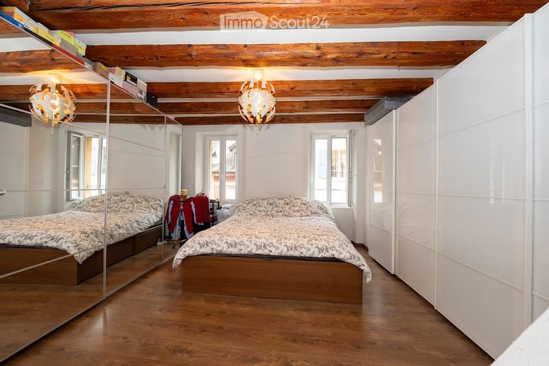 Cozy bedroom with wooden floors, exposed wooden beams, a large mirror, a double bed with patterned bedding, a wardrobe, and two chandeliers.
