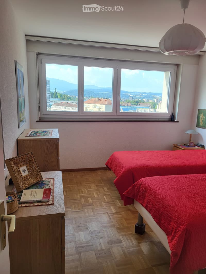 Bright bedroom with wooden flooring, large windows offering a scenic mountain view, a double bed with red bedding, a nightstand with a lamp, a small table with books and a picture frame, and a wicker basket on the floor.