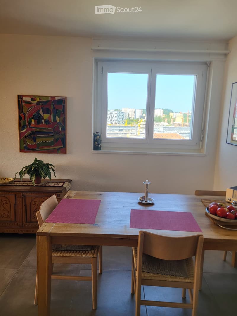 Bright kitchen with a wooden dining table, chairs, a bowl of fruit, a candle, a potted plant, and a colorful painting on the wall. Large windows offer a city view.