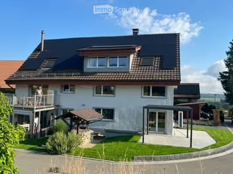 Two-story house with white exterior, dark roof, balcony, and well-maintained garden.