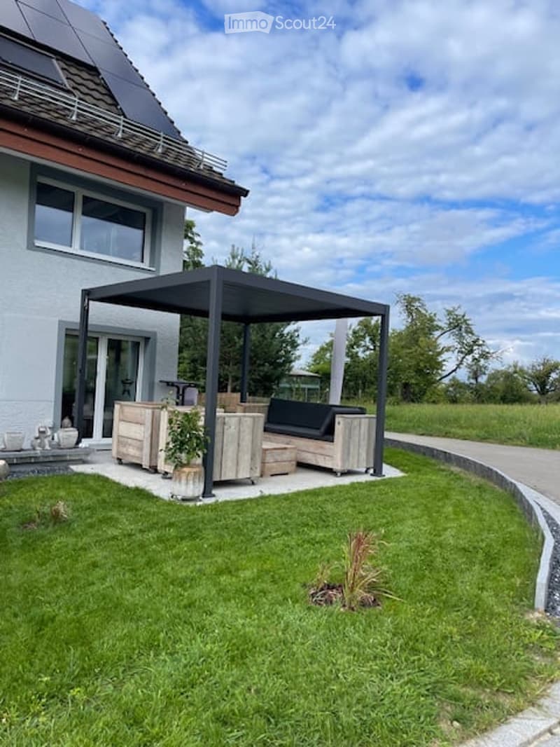 A house exterior with a solar panel on the roof, a wooden bench, potted plant and an open space with outdoor furniture