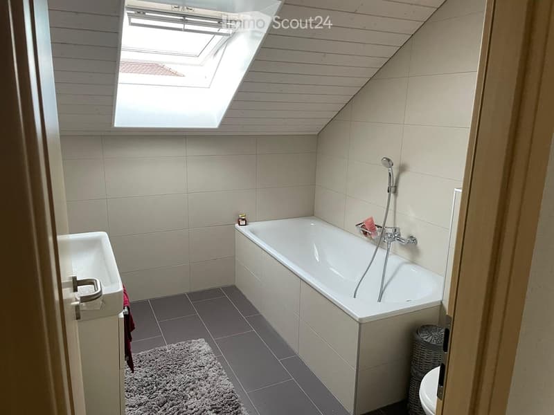 Modern bathroom with a bathtub, showerhead, sink, toilet, and a small wicker basket. The room features a skylight window and a grey tiled floor.