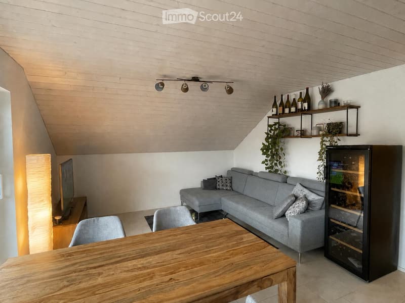 Modern living room with grey sectional sofa, wooden dining table, chairs, wine rack, shelves with bottles and plants, and a floor lamp.