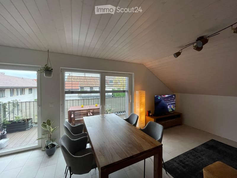 Bright living and dining area with a wooden dining table, chairs, TV, floor lamp, and potted plants. Large sliding glass doors open to a balcony with outdoor furniture.