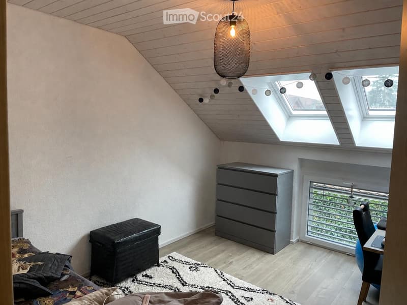 Cozy attic bedroom with wooden slanted ceiling, skylight windows, and modern furniture including a bed, chair, table, chest of drawers, and a stool.