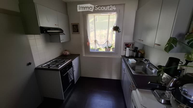 Modern kitchen with white cabinets, gas stove, oven, sink, and coffee maker. Features a window with a curtain and a small plant on the windowsill.