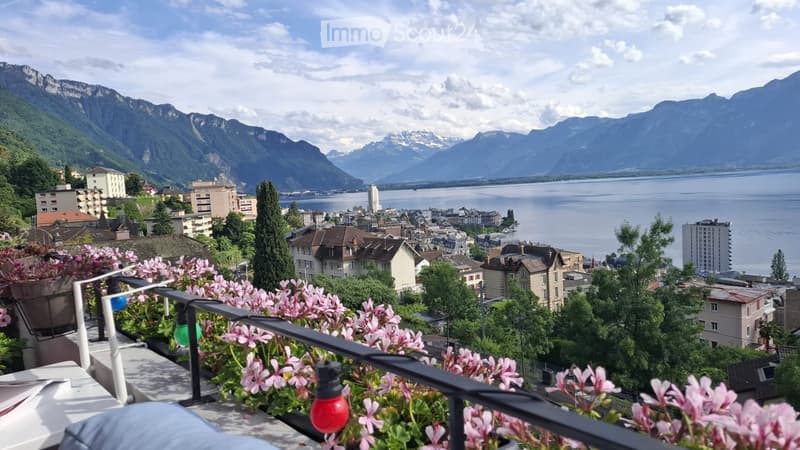 Balcony with pink flowers, overlooking a cityscape with mountains and a lake in the background.