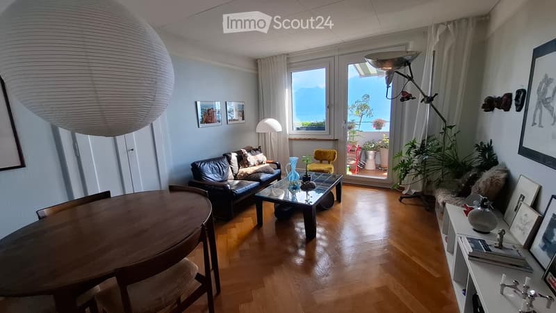 Bright living room with wooden flooring, large windows offering a scenic view, a black leather sofa, a glass coffee table, a wooden dining table, and decorative items including lamps and artwork.