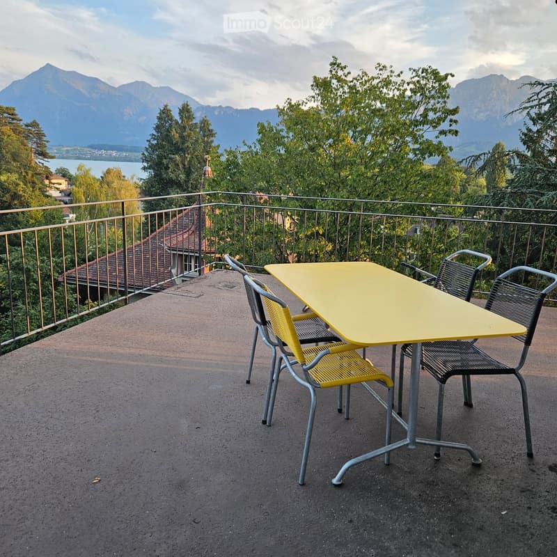 Balcony with a yellow table and two chairs, overlooking a scenic mountain view with trees and a lake.