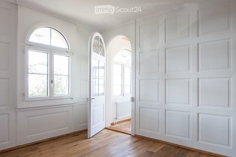 Empty hallway with white walls, wooden flooring, and multiple doors and windows, including arched windows and panelled doors.