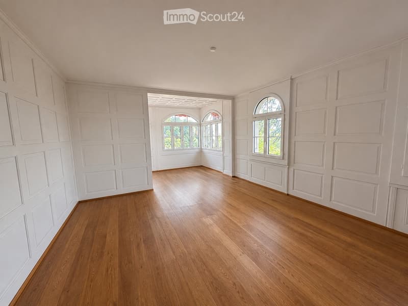 Empty room with wooden flooring and white paneled walls, featuring arched windows and a connecting doorway.