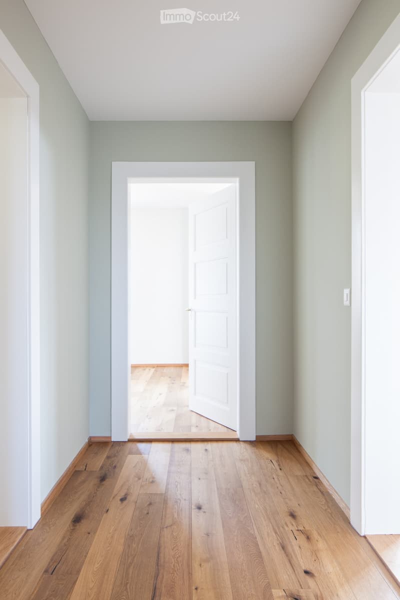 Empty hallway with light green walls, wooden flooring, and an open door leading to another room.