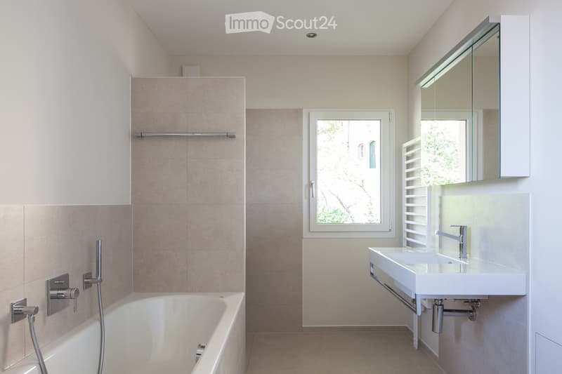 Modern bathroom with a bathtub, sink, mirror, and window offering a view of greenery outside.