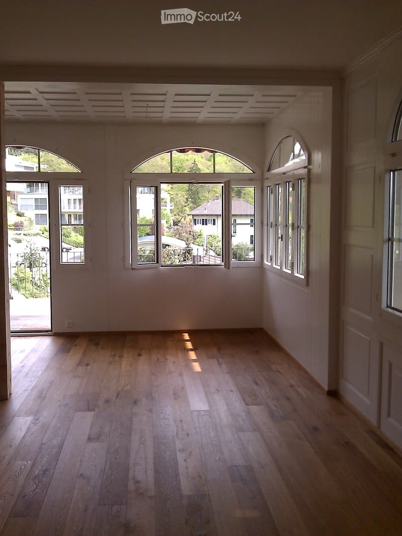 Empty room with light wooden flooring, white walls, and large arched windows offering a view of the outdoors.