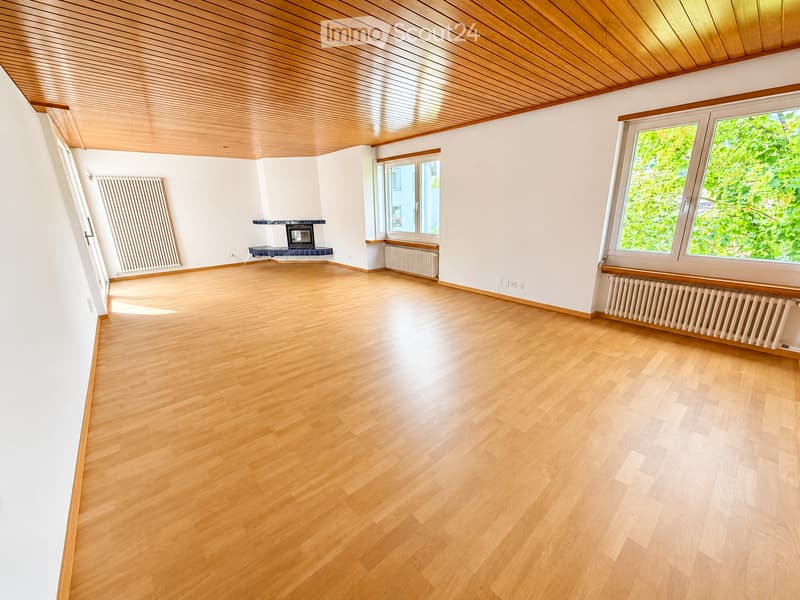 Empty room with wooden flooring, white walls, wooden ceiling, and a fireplace with a blue tiled surround. Radiators are present along the walls, and there are windows with green views outside.