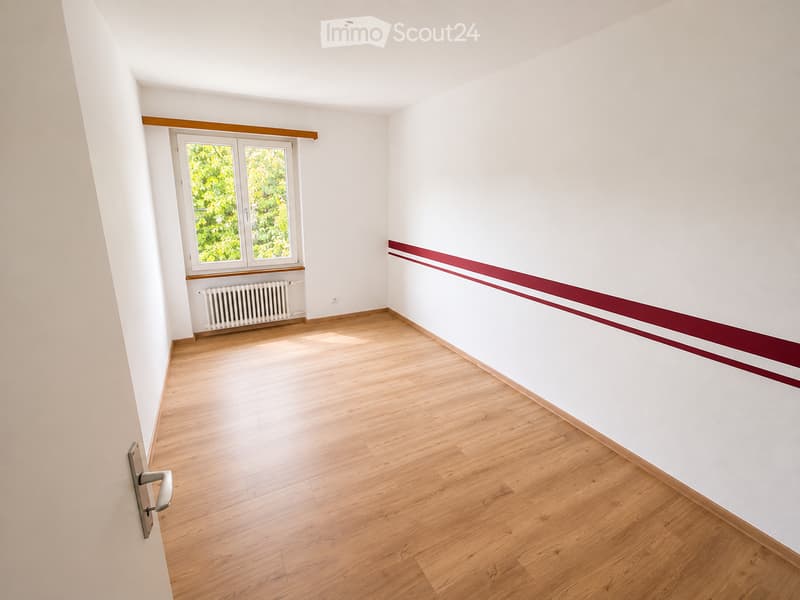 Empty room with wooden flooring, white walls, and a window with a radiator below it. The walls feature a decorative maroon trim near the ceiling.