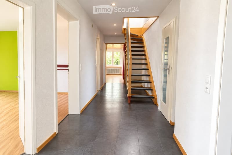 Modern hallway with grey tiled floor, white walls, wooden staircase leading upwards, and several doors opening to adjacent rooms.