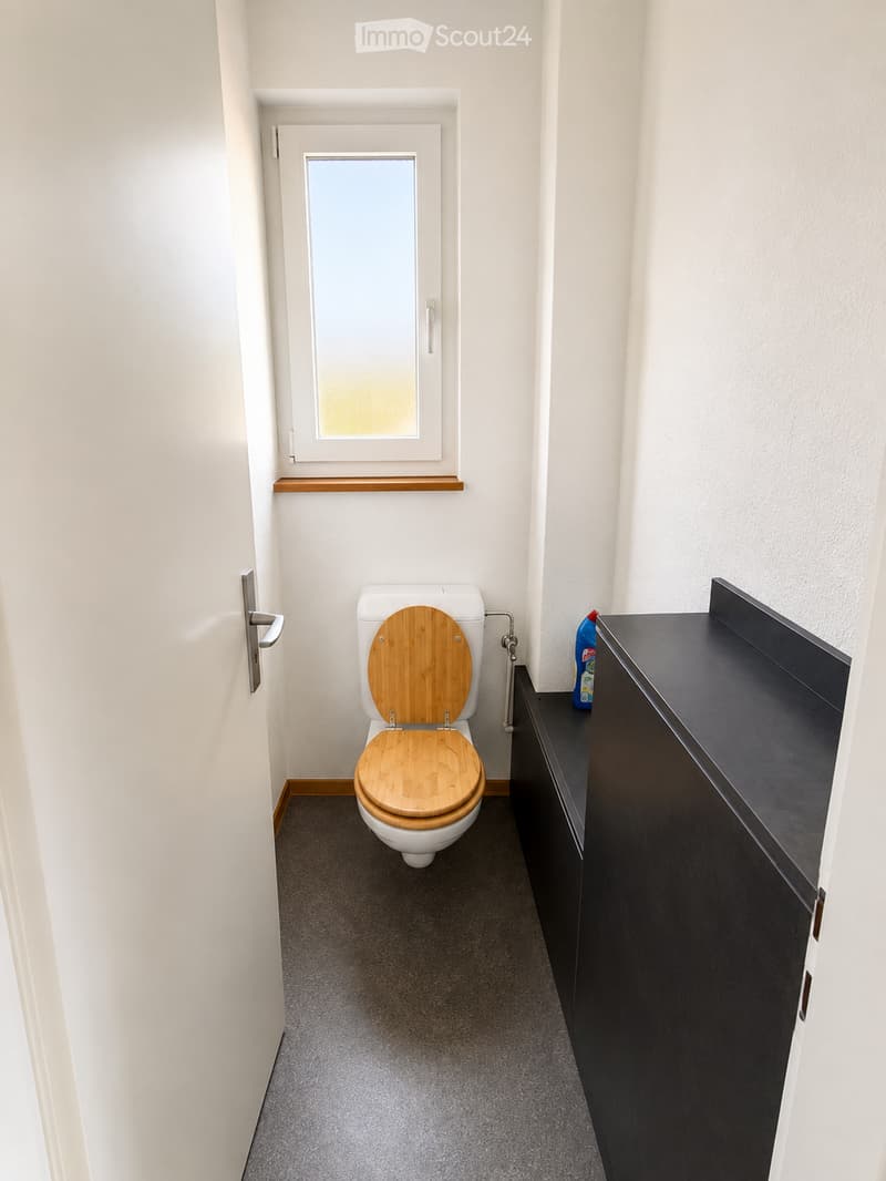 Modern bathroom with a white toilet featuring a wooden seat, a black sink cabinet, and a small window above the toilet.