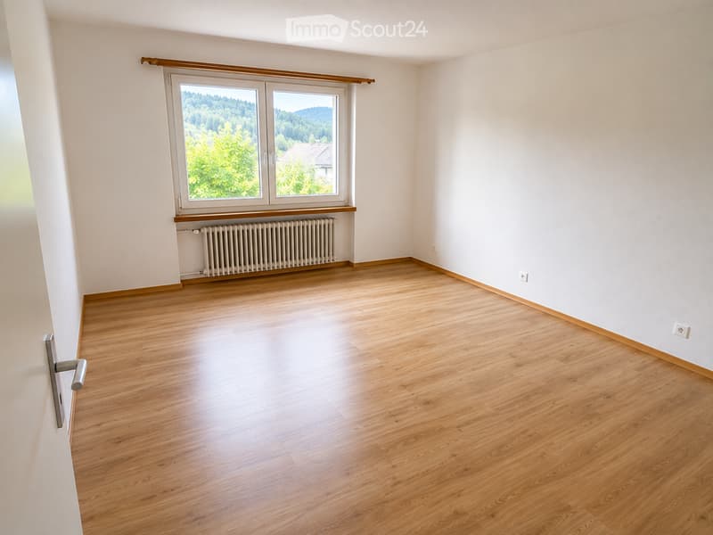 Empty room with wooden flooring, white walls, large window with wooden frame, radiator, and a door with a silver handle.
