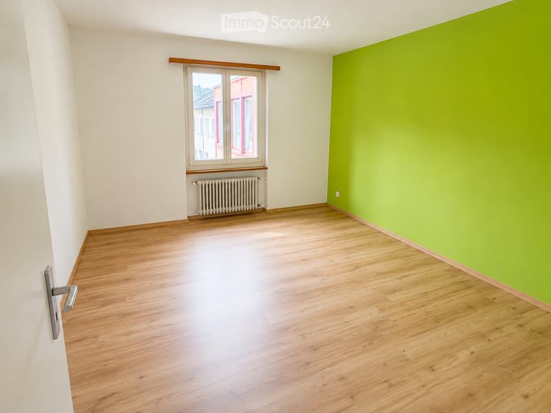 Empty room with wooden flooring, white walls, a green accent wall, a window with a radiator below, and a door with a silver handle.