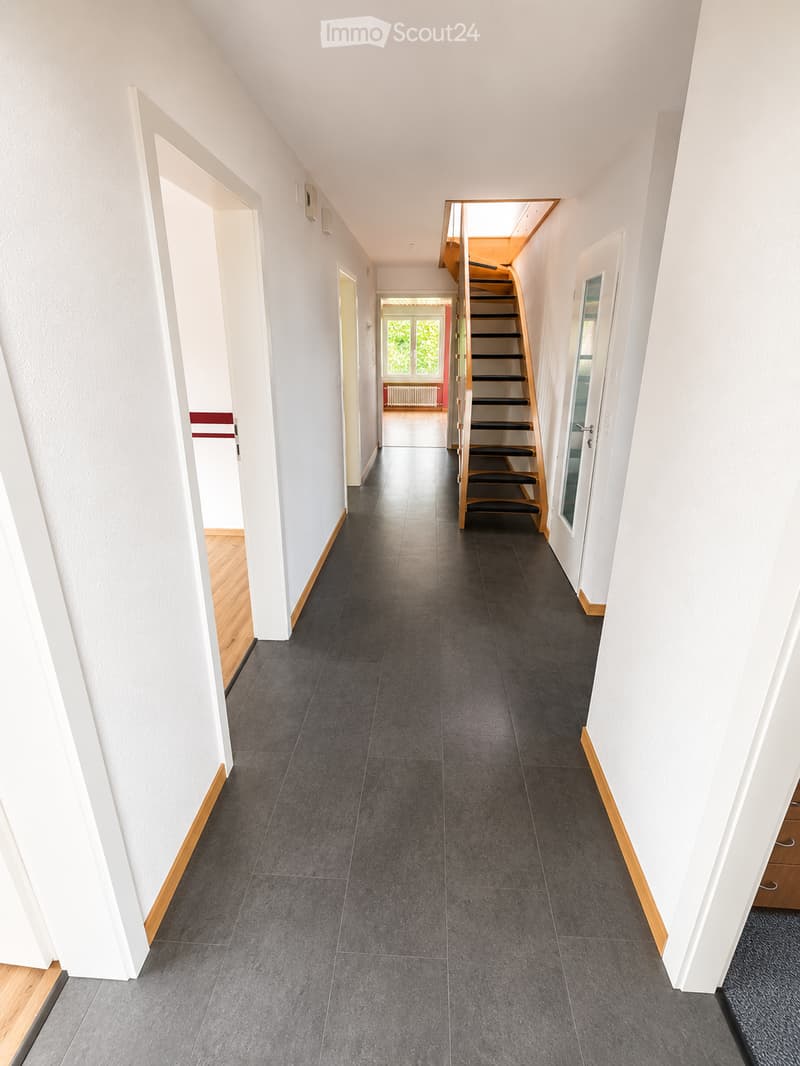 Clean, modern hallway with dark grey tiled flooring, white walls, and wooden trim. Features a staircase with wooden railings and black steps leading upwards, with several doors and a window at the end providing natural light.