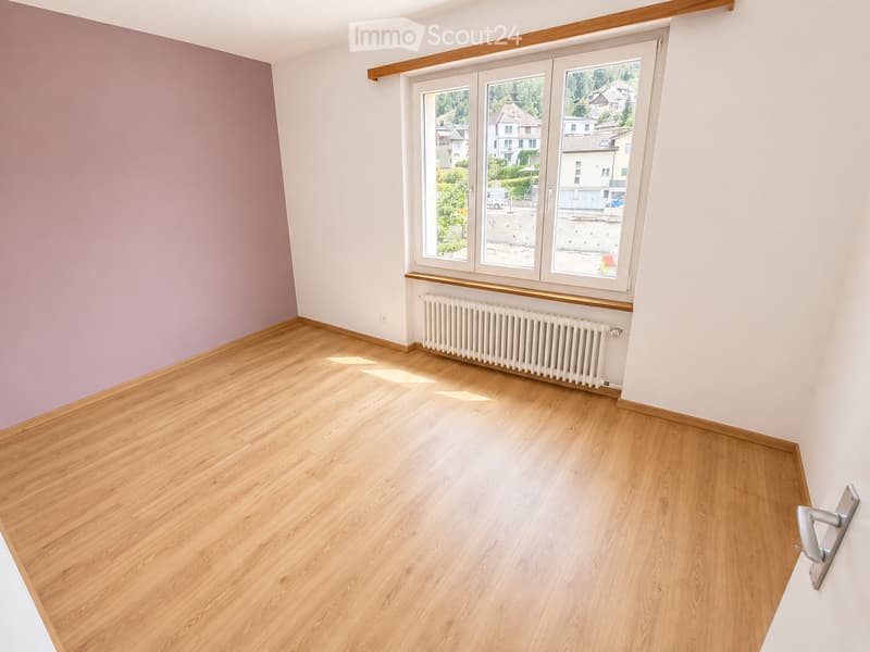 Empty room with wooden flooring, white walls, and large windows offering a view of the outside. A radiator is visible under the windows.