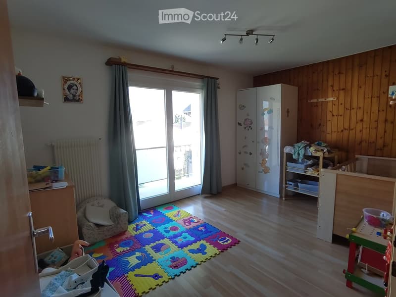 A well-lit room with wooden flooring, a colorful foam play mat, a small sofa, a toy table and chair, a storage cabinet, and a large window with curtains.