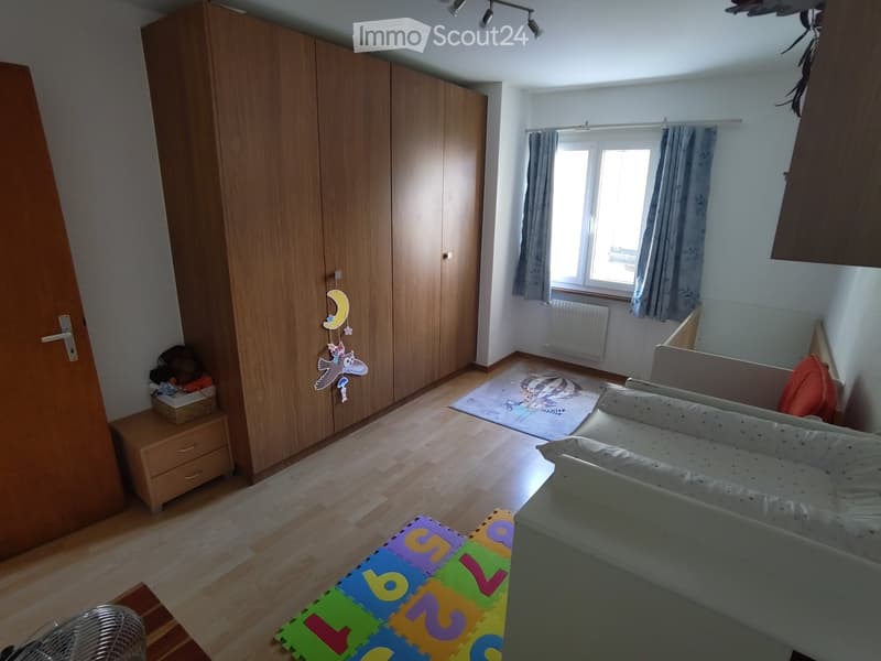 A well-lit children's room with a wooden floor, a large wardrobe, a sofa, a colorful play mat, and a toy box. The room features a window with curtains and a radiator below it.