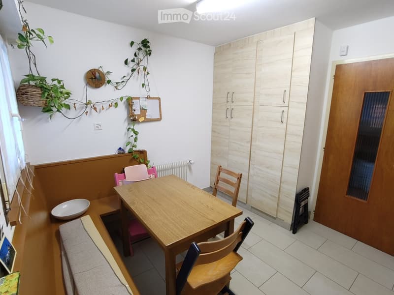A small, well-lit room with a wooden table, chairs, a bench, a bowl, a built-in cupboard, and a hanging plant. The room features a mix of wooden and tiled flooring.