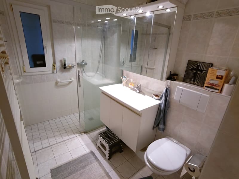 Modern bathroom with white tiles, a large mirror, a sink with a faucet, a toilet, a shower with a glass door, a towel hanging on a rack, a small stool, and a radiator. There is also a window with a curtain and a picture frame on the wall.