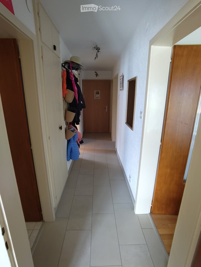 A clean, well-lit hallway with tiled flooring, white walls, and wooden doors. A coat rack with various jackets and hats is visible on the left side.
