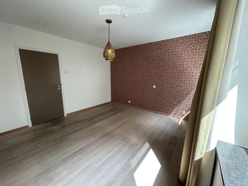 Empty room with wooden flooring, white walls, and patterned wallpaper on one wall. Features a hanging light fixture and a closed door.