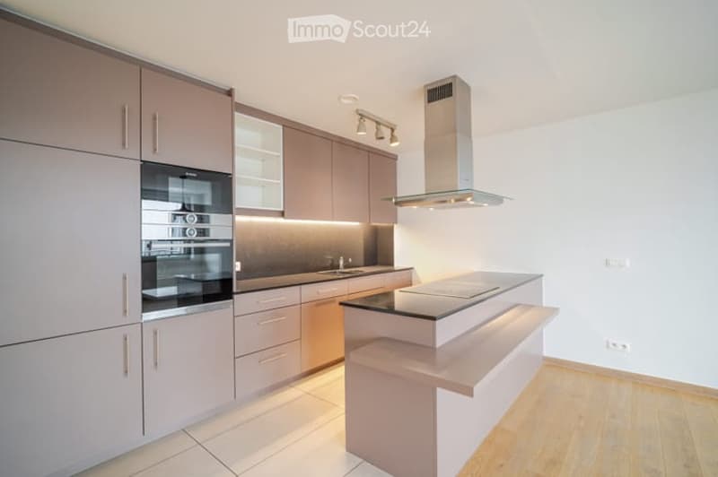 Modern kitchen with light-colored cabinetry, stainless steel oven, island, and range hood. Clean, bright, and well-lit with wooden flooring.