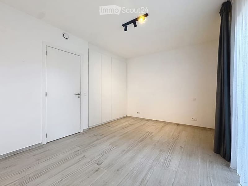 Empty room with light wooden flooring, white walls, a white door, and a window with dark curtains. The room has a modern, minimalist design with recessed lighting.