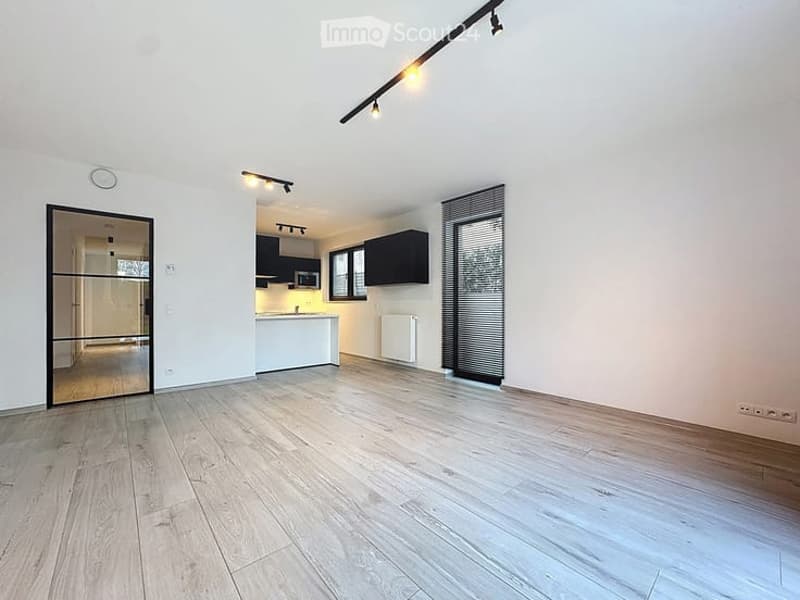 Open-plan unfurnished room with light wooden flooring, white walls, and a modern kitchen area in the background. The kitchen features black cabinets, a white countertop, and built-in appliances.