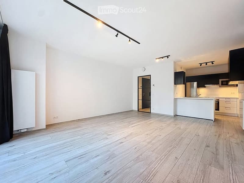Empty room with light wooden flooring, white walls, and a modern kitchen area in the background. No furniture or personal items are present.